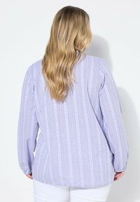 Camisa de manga larga lavanda con patrones florales blancos verticales, de corte relajado y con puños elásticos, mostrada desde la parte posterior.