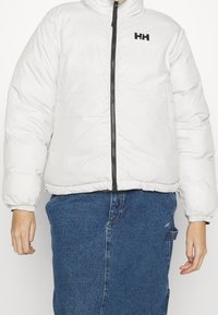 Personne portant une doudoune blanche avec fermeture éclair à l'avant et logo HH noir, associée à une jupe en denim bleu taille haute avec poche et passants.