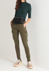 Olijfkleurige cargobroek met zijzakken, gecombineerd met een kortebloes in donkergroen en een zwarte heuptas. Beige en roze sneakers met een patroon.