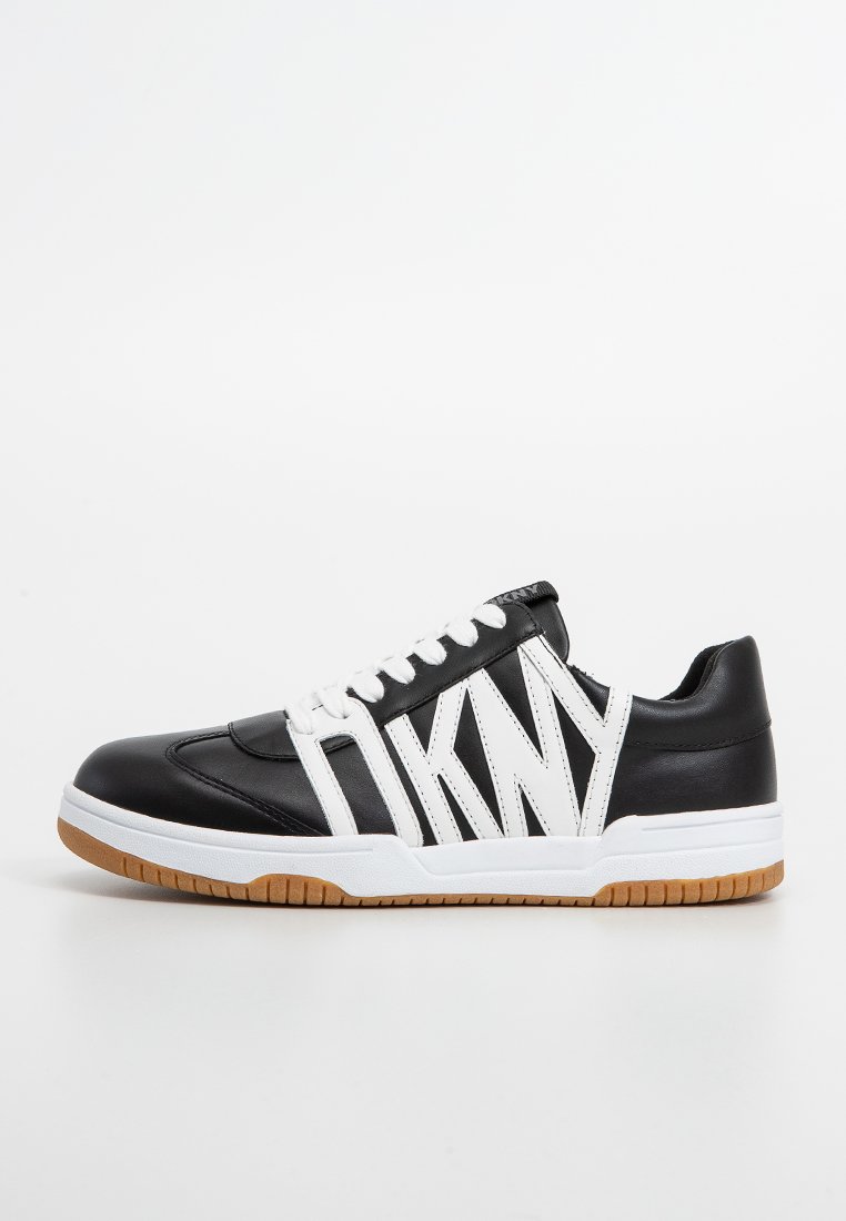 Zapatilla deportiva en blanco y negro con una parte superior de cuero suave, que presenta franjas blancas llamativas, una suela blanca y una suela de goma color caramelo.