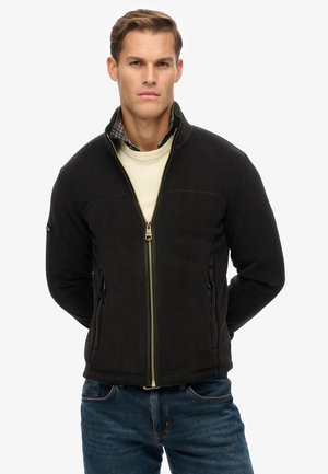 Zwarte fleece jack met ritssluiting, zijzakken en een groene accentrits. Draagt over een crèmekleurige trui en een geruite blouse.