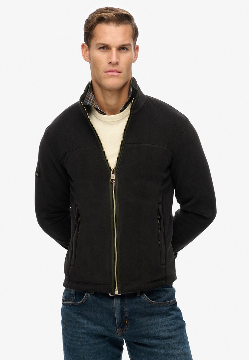 Zwarte fleece jack met ritssluiting, zijzakken en een groene accentrits. Draagt over een crèmekleurige trui en een geruite blouse.