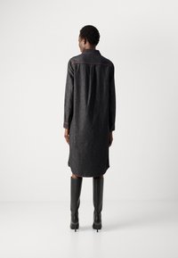 Kvinne i en mørk denim langermet skjortekjole med røde sømmer og sorte høye støvler til knærne, stående mot en enkel hvit bakgrunn.