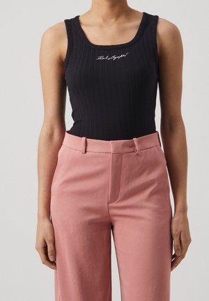 Žena v černém bezrukávovém ribbed topu s bílým nápisem a růžových kalhotách s vysokým pasem, stojící před jednobarevným pozadím.