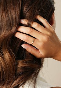 Goldring mit einem glatten, geschwungenen Design, der an einer Hand getragen wird, die wellige, dunkelbraune Haare berührt. Weiche, natürliche Nagelfarbe sichtbar.