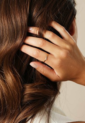 Guld ring med et glat, buet design blevet båret på en hånd, der rører bølget, mørkebrunt hår. Bløde, naturlige neglefaver er synlige.