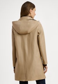 Beige utomhuskappa med huva, rak skärning, långa ärmar och en baktill slits för rörlighet. Har slät bomullsblandad tyg.