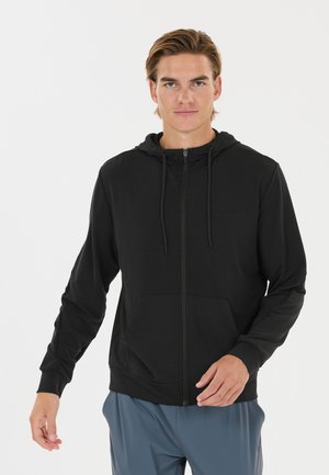 Junger Mann, der einen schwarzen Kapuzenpullover mit Reißverschluss und Kordeln sowie blaue Hosen trägt, steht vor einem schlichten weißen Hintergrund.