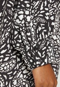 Blusa com padrão em preto e branco, apresentando um tecido texturizado, mangas bufantes e punhos com botão, decorados com três botões escuros.