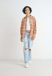 Rutet skjorte i orange og beige nuancer, parret med en hvid T-shirt, slidte lyseblå jeans og hvide sneakers. Enkel design, afslappet pasform.