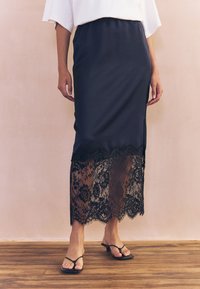 Granatowa ołówkowa spódnica z koronkową nałożoną warstwą przy dolnym brzegu, zdobiona kwiatowymi wzorami. Posiada boczny rozcięcie i jest stylizowana z prostymi czarnymi sandałami.