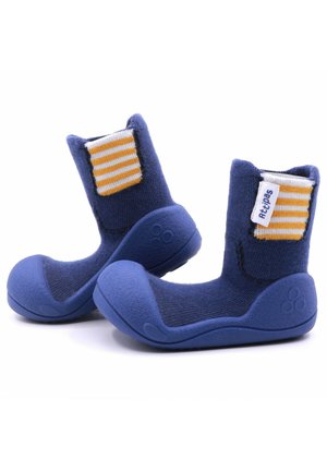 Botitas azules hechas de tela suave con una suela de goma texturizada. Presentan un acento a rayas amarillas y blancas en la parte trasera.