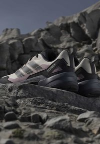Ζευγάρι μαύρα και λευκά παπούτσια δρόμου Adidas Terrex που ξεκουράζονται σε πετρώδες έδαφος με τραχιά βουνά στο φόντο.