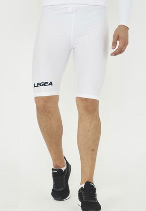 Uomo che indossa pantaloncini sportivi bianchi aderenti con logo "LEGEA" e scarpe da corsa nere che cammina in avanti su sfondo bianco.