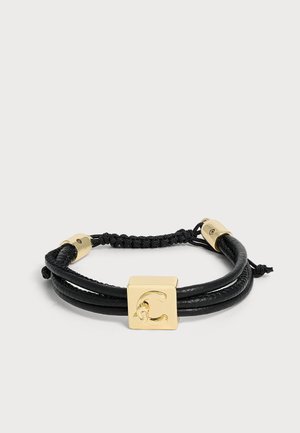 Pulsera de cuero negro con detalles metálicos dorados y un colgante cuadrado central grabado con la letra C, cierre de cordón ajustable.
