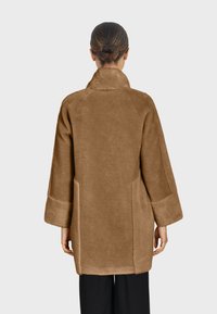 Manteau en fausse fourrure marron avec un col, des manches trois-quarts et des poches latérales. Présente une texture douce et une coupe décontractée. Vue arrière montrée.