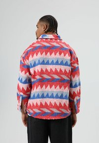 Buntes Wollhemd mit einem geometrischen Muster in Rot, Blau und Beige, mit Kragen und lockerem Schnitt, sichtbare Seitenschlitze.