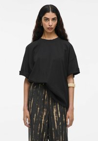 Juoda per didelė t-shirt su sukamu rankovėmis, derinama su laisvomis, raštuotomis tamsiomis kelnėmis su rudomis vertikaliomis juostelėmis. Sklandžios audinio tekstūros.