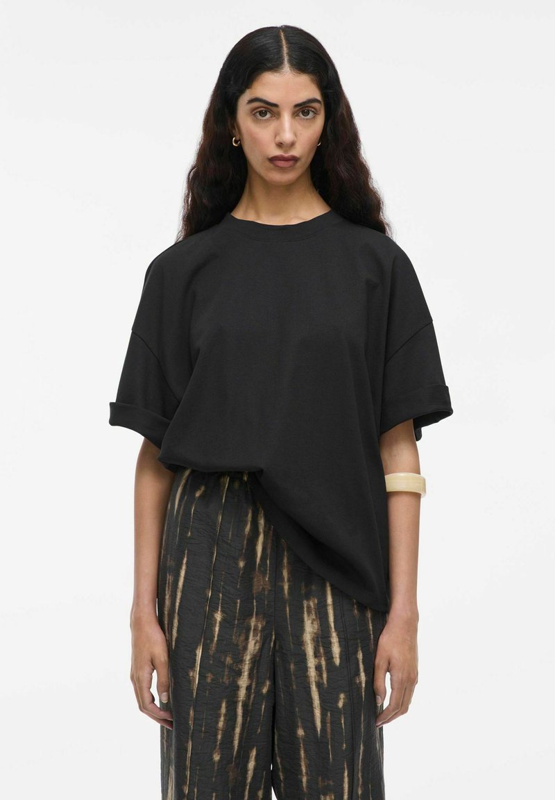 Juoda per didelė t-shirt su sukamu rankovėmis, derinama su laisvomis, raštuotomis tamsiomis kelnėmis su rudomis vertikaliomis juostelėmis. Sklandžios audinio tekstūros.