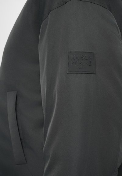 Chaqueta negra con una textura suave, que presenta un parche en el pecho con el texto "MAISON KITSUNÉ PARIS" y un diseño de bolsillo lateral.