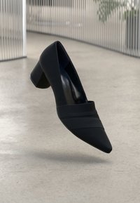 Black high-heeled shoe with a pointed toe and angular design. Made of textured material with two horizontal stripes across the vamp.