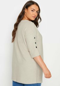Licht beige oversized top met halve mouwen. Bevat drie zwarte knopen op de mouwen en is gemaakt van een zachte, gestructureerde stof.