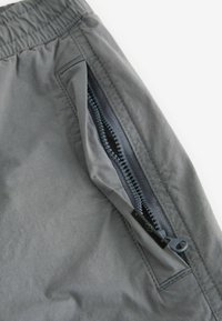 Tissu gris avec une ceinture élastique montrant une fermeture éclair partiellement ouverte sur une poche latérale d'un pantalon léger.
