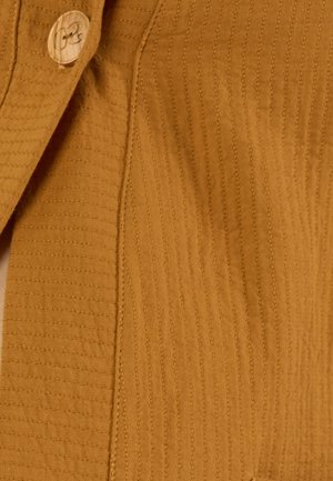 Close-up van mosterdgele textielstof met verticale gestikte lijnen en een ronde beige knoop met een motief.