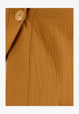 Close-up van mosterdgele textielstof met verticale gestikte lijnen en een ronde beige knoop met een motief.