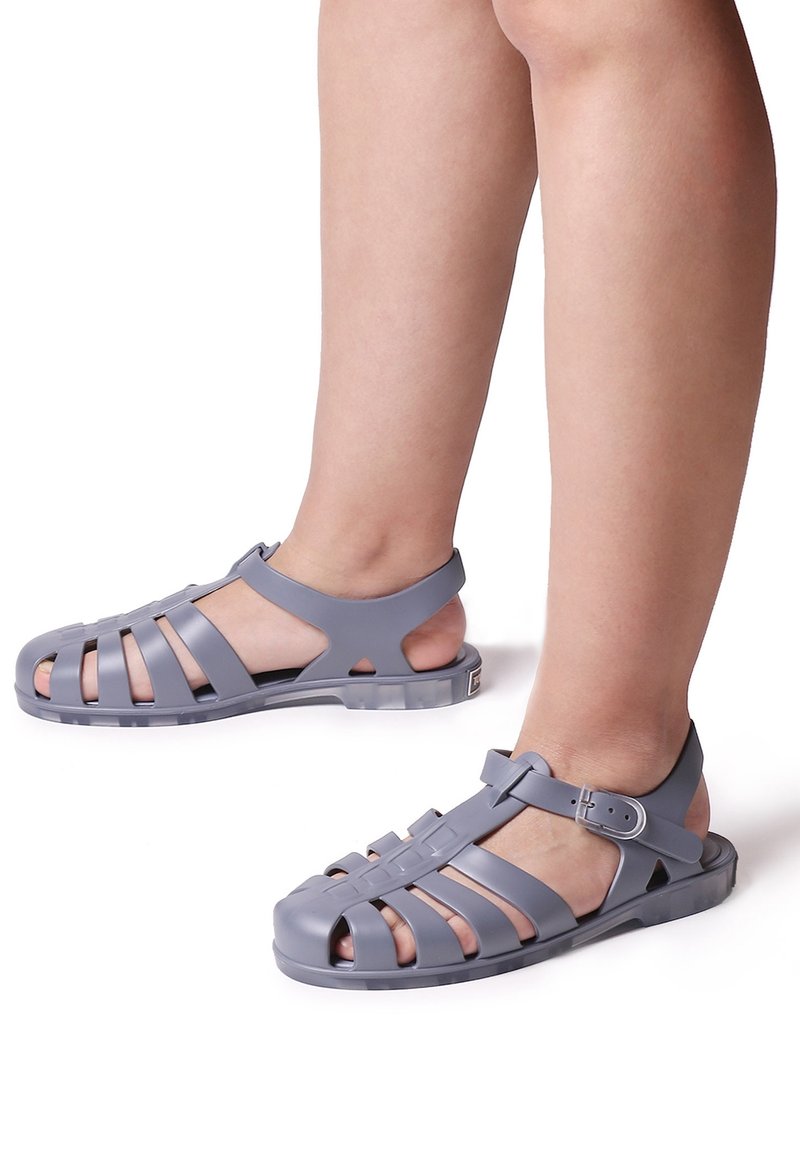 Jambes d'enfant portant des sandales en plastique gris à bout fermé avec des sangles réglables à boucle sur fond blanc.