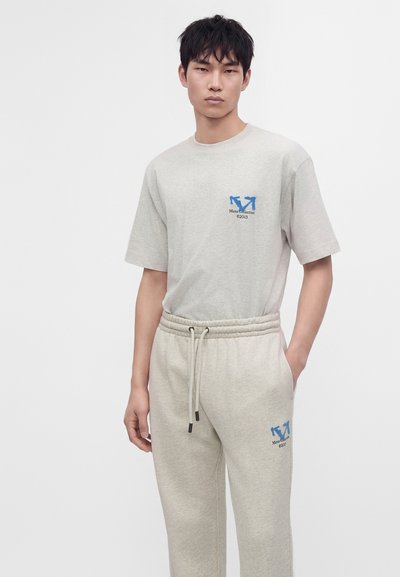 Jeune homme portant un t-shirt beige clair et un pantalon de survêtement avec des logos fléchés bleus et du texte, debout, une main dans la poche, sur fond blanc.