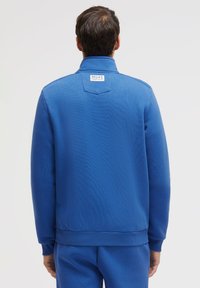 Blue fleece jacket with a high collar, textured surface, and ribbed cuffs. Features a tag on the back neck area, no visible fastenings.