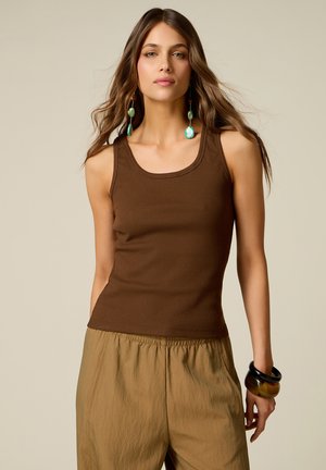 Donna con capelli lunghi e mossi che indossa una canottiera marrone, pantaloni larghi color kaki, grandi orecchini verdi e braccialetti spessi al polso.