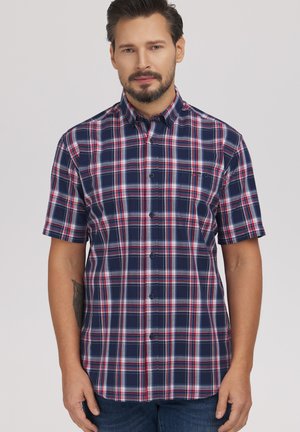 Mężczyzna o krótkich ciemnych włosach i brodzie, ubrany w granatową, czerwoną i białą kratowaną koszulę na krótki rękaw, na jednolitym tle.