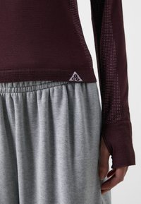 Bordeaux långärmad topp med texturerade ärmar, kombinerad med en grå plisserad kjol. Har en triangulär logotyp på midjebandet.
