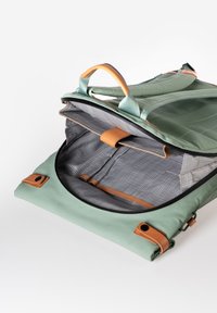 Zaino morbido verde con dettagli in pelle marrone. Presenta un ampio interno, foderato con un motivo a rete grigia, e doppi manici superiori per il trasporto.