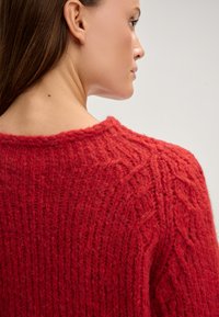 Maglione rosso lavorato a maglia con un motivo testurizzato, che mostra dettagli a coste e un collo rotondo. Il tessuto appare spesso e accogliente.