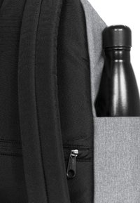 Mochila con un lado de tela negra y un frente de tela gris, que presenta un zip plateado visible y una botella de agua negra en el bolsillo lateral.