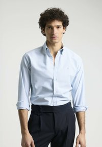 Camisa de dress raya azul claro con cuello abotonado, mangas largas remangadas en los puños, combinada con pantalones oscuros de cintura alta con detalles de botones.