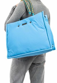 Lichtblauwe totebag met gequilt ontwerp, gestructureerde stof en contrasterende gevlochten schouderbanden in blauw en geel. Twee zwarte knopen aan de voorkant.