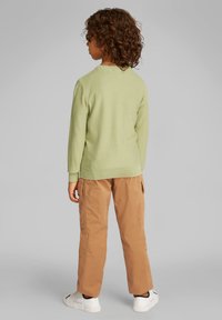 Bambino con capelli ricci che indossa un maglione verde chiaro, pantaloni cargo beige e sneakers bianche, in piedi di spalle su uno sfondo grigio semplice.