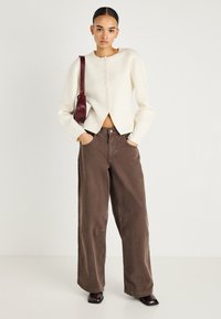 Krēmkrāsas vilnas maisījuma kardigans ar apaļu kakla izgriezumu un priekšpusē pogātu aizdari, kopā ar brūnām plaša piegriezuma augstvidukļa džinsu biksēm un bordo plecu somu.