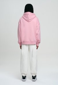 Sweat à capuche rose oversize avec une grande capuche, poignets côtelés, porté avec un pantalon de survêtement blanc et des chaussures de sport noires. Design épuré et minimaliste.