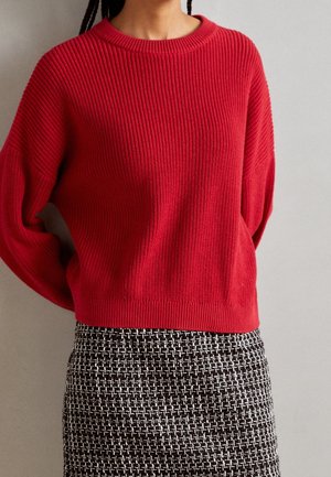 Donna che indossa un maglione a coste rosso e una gonna a quadri bianco e nero, in piedi davanti a uno sfondo neutro.