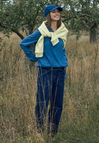 Frau lächelt im Freien im hohen Gras, trägt eine blaue Mütze, ein blaues Hemd, einen gelben Pullover über den Schultern und dunkelblaue Jeans.