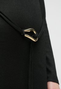 Čierny textilný odev s tenkým čiernym šnúrkou upevneným lesklou zlatou trojuholníkovou prackou v páse.