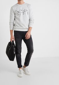 Man i ljusgrå Hugo Boss sweatshirt, svarta jeans, vita sneakers, som håller en svart portfölj mot en plain vit bakgrund.