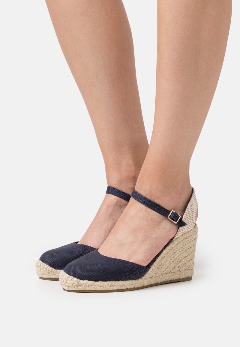 Espadrilles compensées en toile bleu marine avec détails en jute tressé, bride à la cheville et bout rond. La tige texturée et le talon en fibre naturelle créent un contraste.