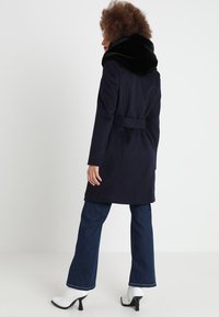Marineblaue Jacke mit schwarzem Kunstpelzkragen, tailliertem Bund und geradem Schnitt. Getragen über ausgestellten Jeans und weißen Stiefeletten. Einfarbiger Hintergrund.