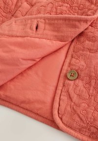 Korallszínű szövet virágmintás hímzéssel, gombbal és gomblyukkal, részben összefordítva, hogy látható legyen a sima belső bélés.